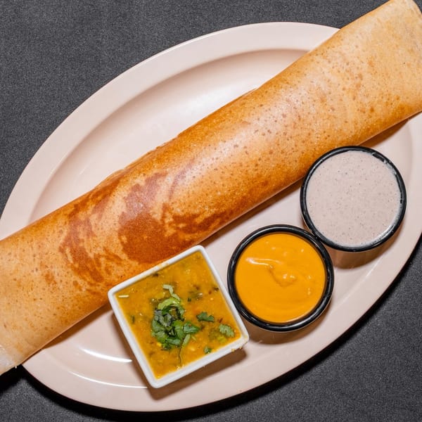 A golden-brown dosa roll on a light pink plate, served with three small bowls of sauces.