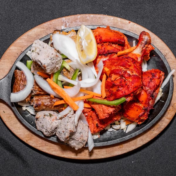 Sizzling platter with tandoori chicken, seekh kebabs, and malai boti, garnished with onions, carrots, and lemon.