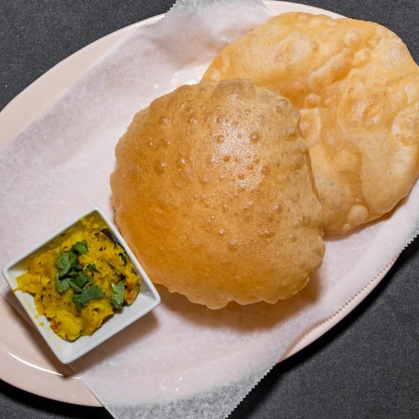 Two golden-brown puri breads and a small dish of yellow potato curry with cilantro on a plate.