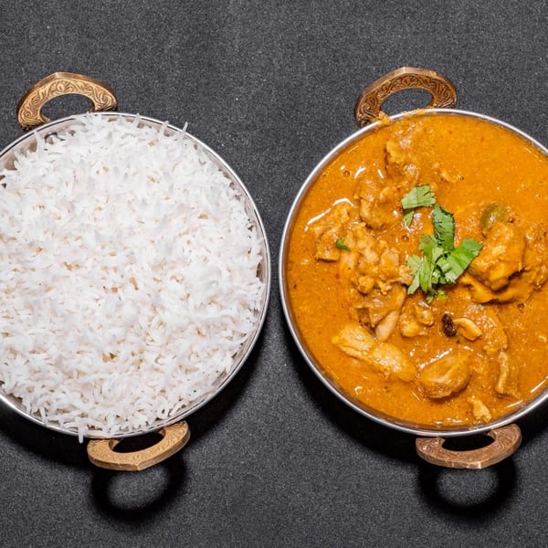 Two metal bowls, one with white rice and the other with chicken curry, on a dark surface.