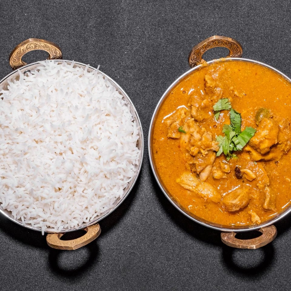 Two metal bowls, one with white rice and the other with chicken curry, on a dark surface.